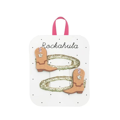 A pair of western-themed hair clips on a white display card with a pink ribbon loop. The clips feature tan faux leather cowboy boots with rhinestone embellishments and glittery gold rope accents.