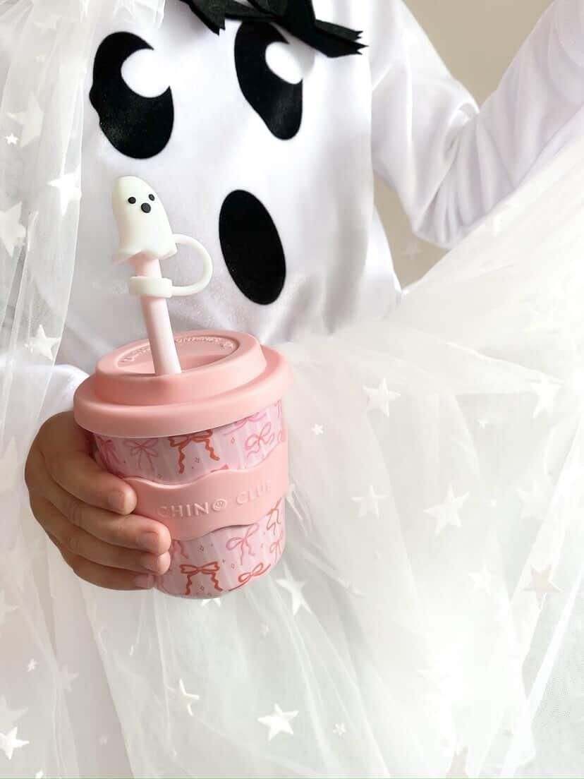 A child wearing a white ghost costume with black eyes and a black bow tie, holding a pink reusable coffee cup with a ghost-shaped straw.