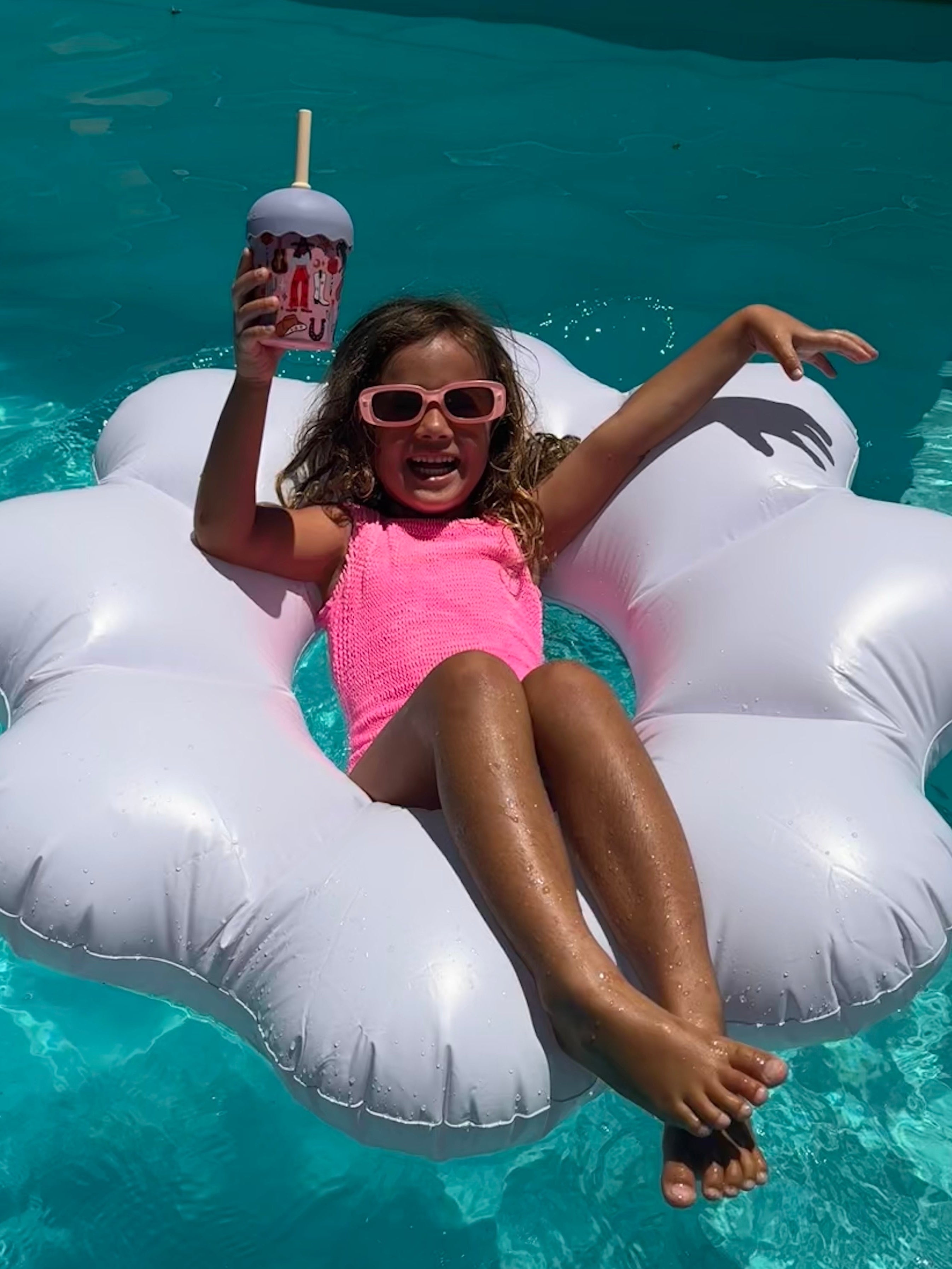Child on a cloud-shaped inflatable pool float holding a smoothie cup drink in a pool with straw in cowgirl design