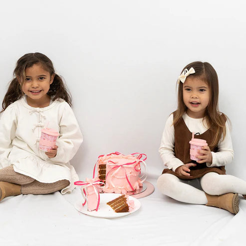 Two young girls with pink bow babychino cups and a cake on a white background