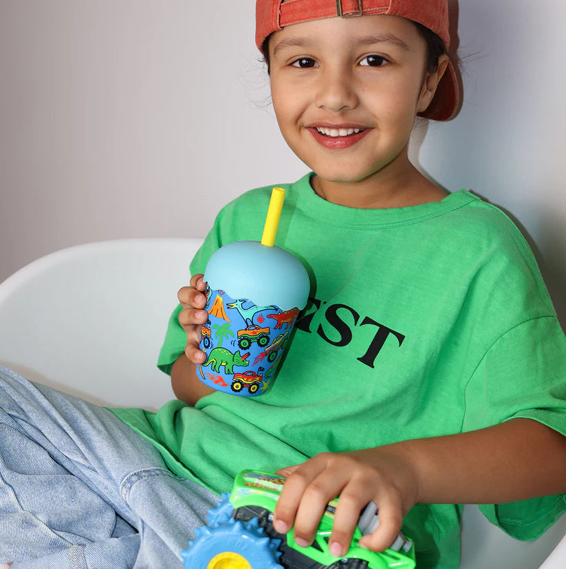 A smiling young boy wearing a green t-shirt and a red cap holds a light blue cup with a yellow straw decorated with colorful toy vehicles. He is sitting in a white chair against a neutral background.