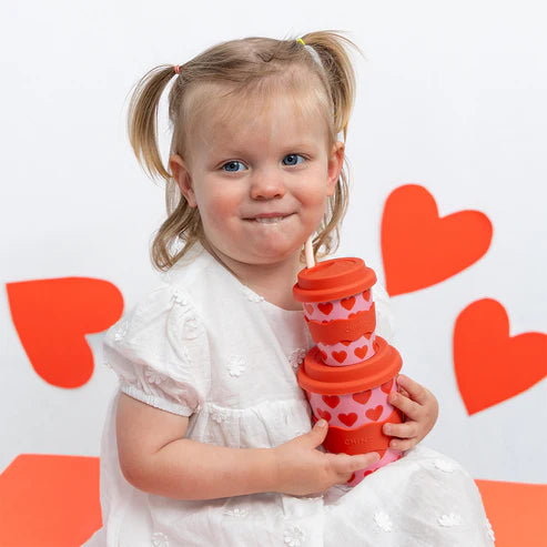 Child holding a red cup with heart patterns against a white background with red hearts. 4oz and matching 8oz babyccino cup
