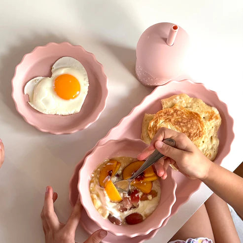 Pink silicone plate and bowl set with breakfast items, including eggs and pancakes on a white surface.