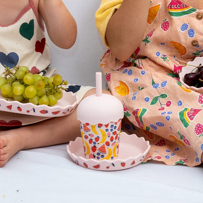 Colorful children's cup with fruit design on a white surface, surrounded by children in patterned outfits.